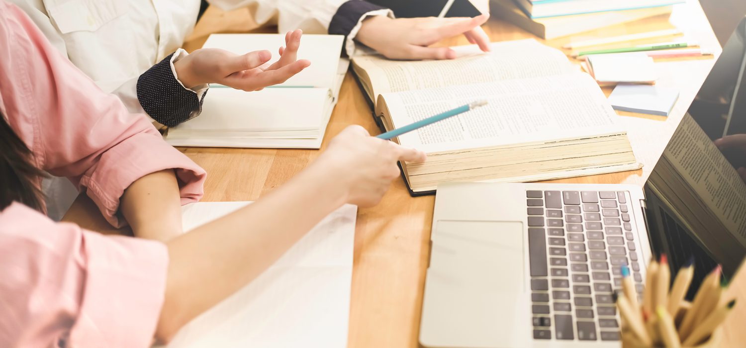 High,School,Or,College,Using,Laptop,While,Sitting,At,Table. High school or college using laptop while sitting at table. Group students studying and reading with books in library. Students helps friend catching up and learning tutoring. education concepts